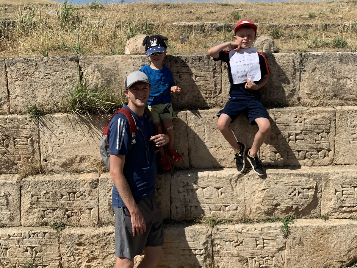 Jamie Watt and two of his sons at a stone wall carved with cuneiform, one boy holding up a printed cuneiform chart